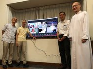 The men take a group photo with a screen which includes those in the Zoom reunion for the 40th anniversary.