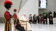Pope Francis seated on a podium flanked by two Swiss guards, view from the right