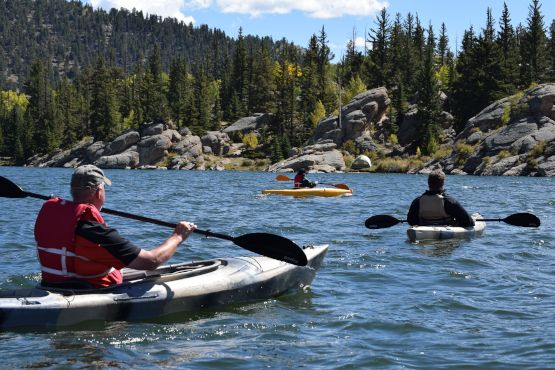 Trois kayakistes s'approchent dans un lac à un rivage rocheux parsemé de conifères.