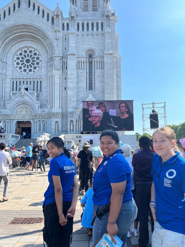 Trois bénévoles devant la basilique Sainte-Anne-de-Beaupré. Un écran géant à la droite diffuse un chant par la chorale.