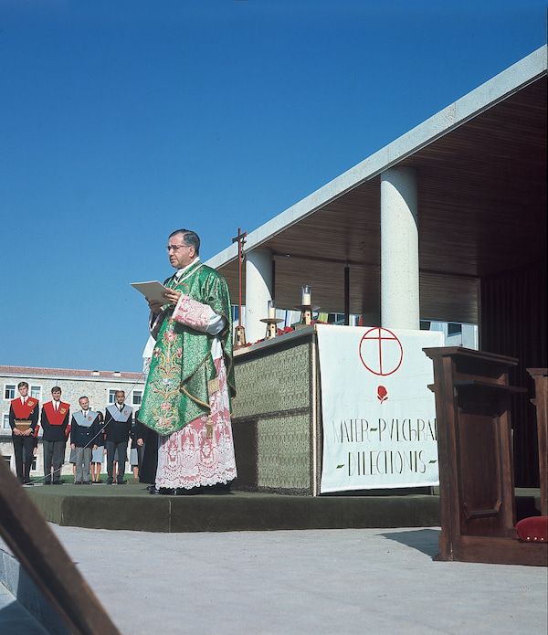 En plein-air, Saint Josémaria devant l'autel, s'apprête à lire. En retrait, sur sa droite, de jeunes hommes debout sont attentifs.