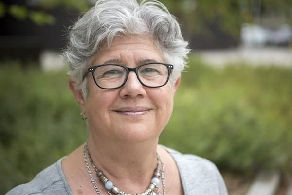 A friendly headshot of Diane, a woman with short gray hair and glasses, smiling. She wears a necklace; greenery blurs softly behind her.
