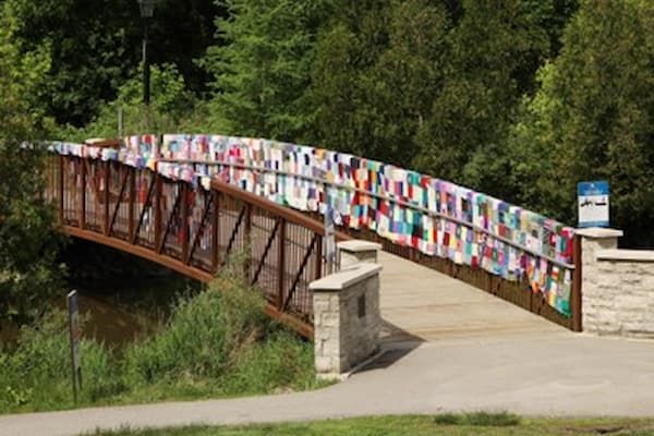 A pedestrian bridge, adorned with colorful knitted squares (yarn bombing), spans a river amidst lush green trees.