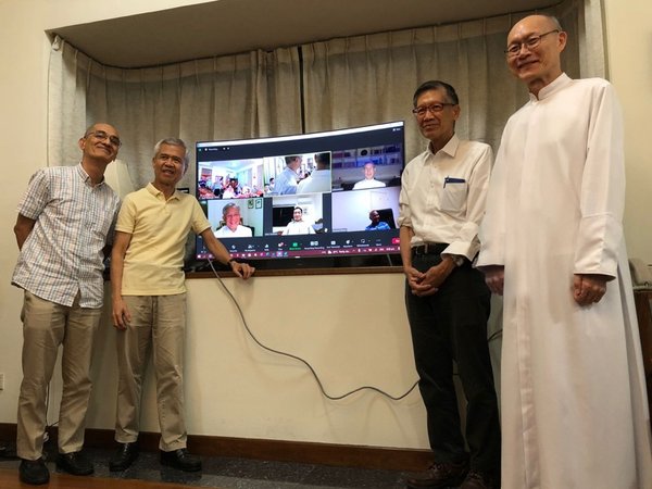 The men take a group photo with a screen which includes those in the Zoom reunion for the 40th anniversary.